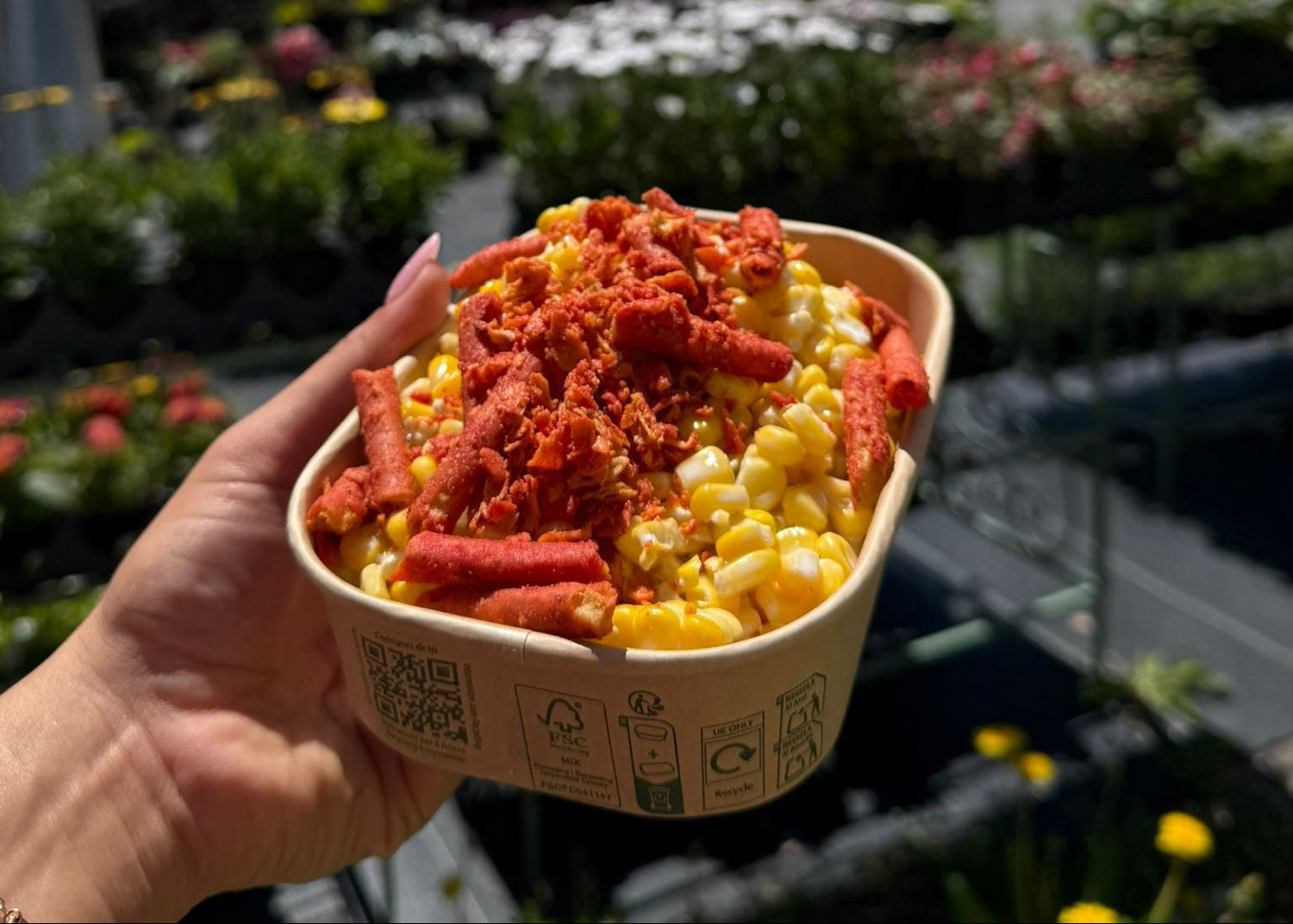 A hand holds a paper bowl filled with corn, red seasoning, and hot dogs outdoors at a plant store, with a white tent and flowers blurred in the background.