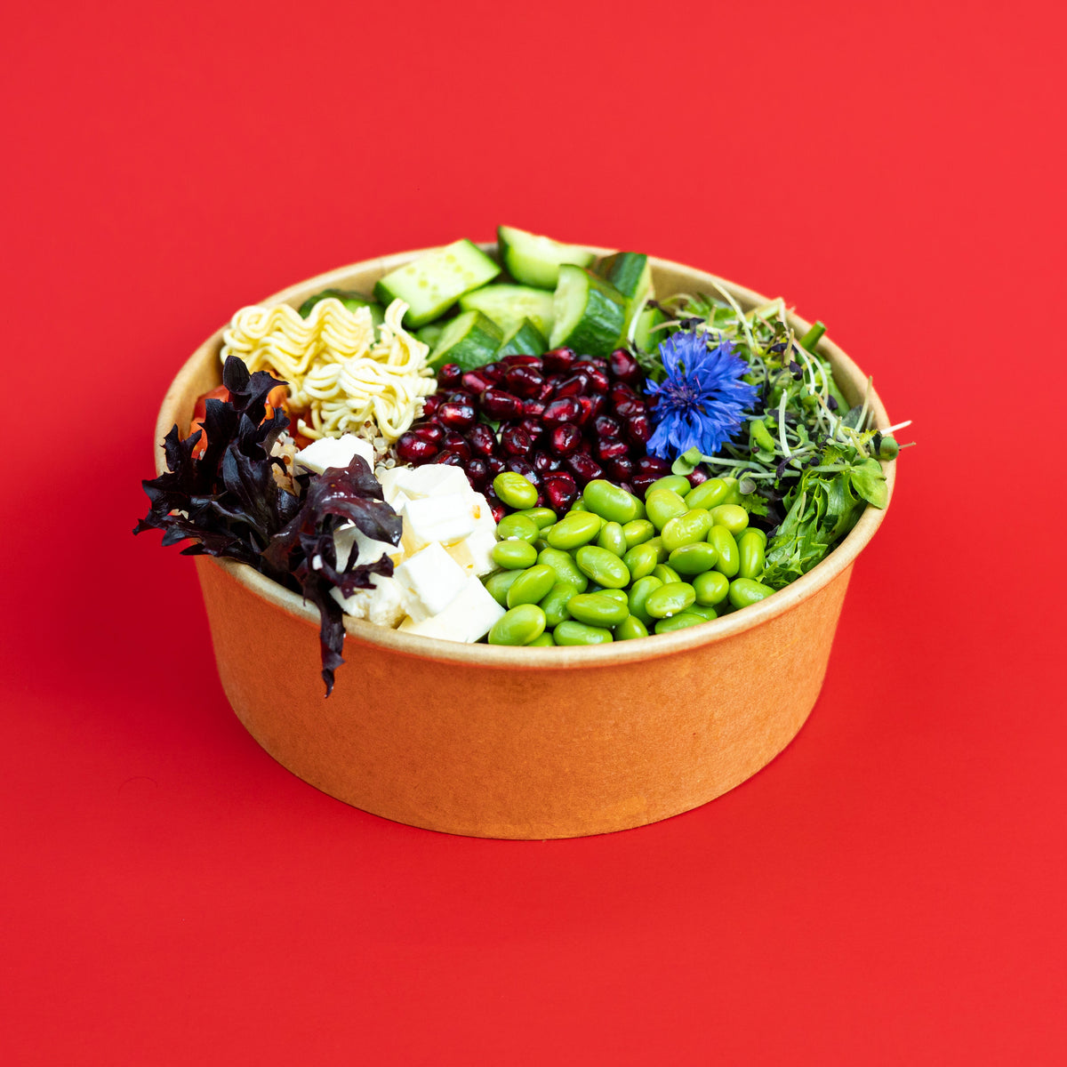A paper bowl filled with assorted salad ingredients, including green edamame, cucumber, mixed greens, red beans, white cheese, and dark leafy greens, set against a solid red background.