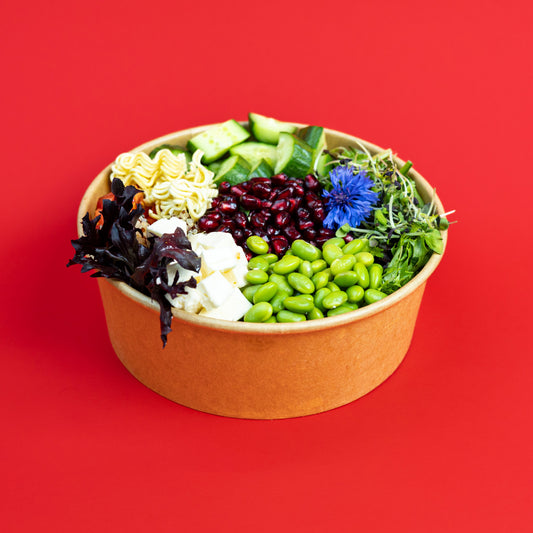 A paper bowl filled with assorted salad ingredients, including green edamame, cucumber, mixed greens, red beans, white cheese, and dark leafy greens, set against a solid red background.