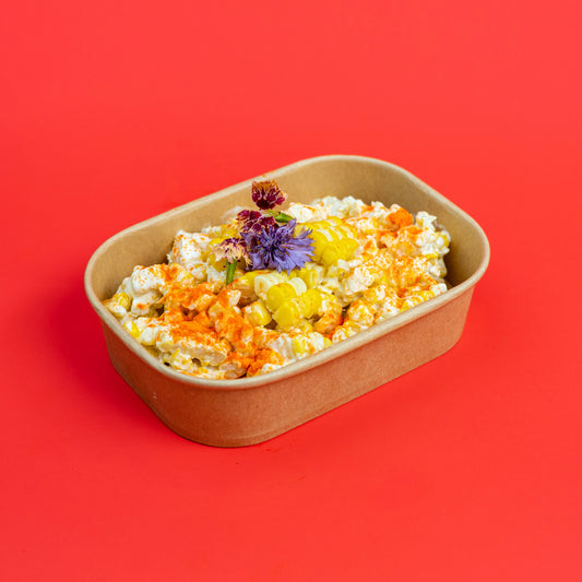 A paperboard rectangular container filled with popcorn and topped with orange zest, yellow fruit pieces, and a small purple flower, set against a solid red background.