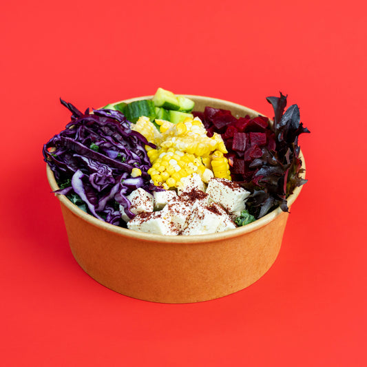 A paper bowl filled with a colorful salad, featuring purple cabbage, red cabbage, corn, cucumber, and white beans, set against a solid red background.