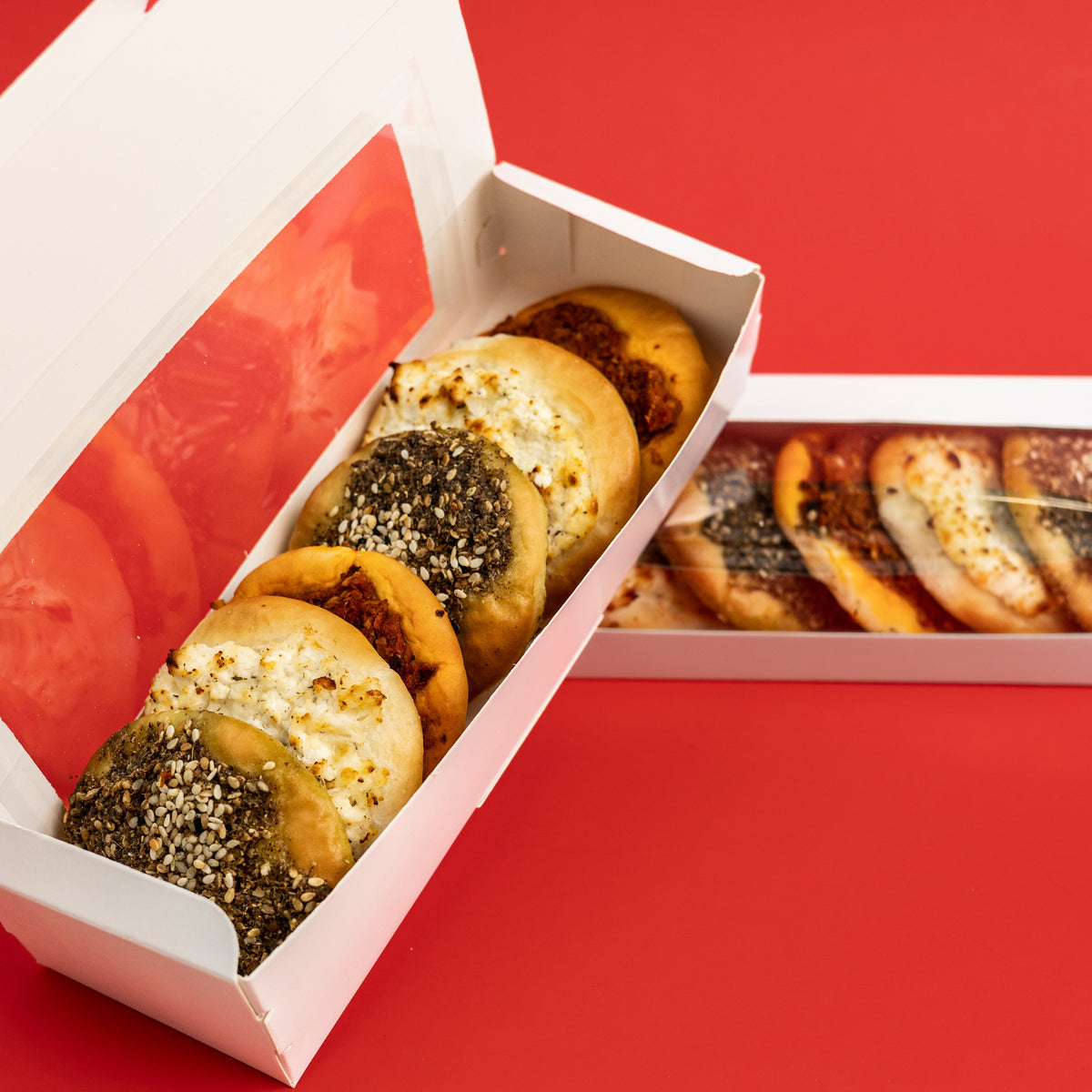 Two white cardboard bakery boxes containing assorted bagels and round baked goods, photographed on a solid red background. One box is open, showing the front bagels, and the other lies beside it showing the back side with a clear window.