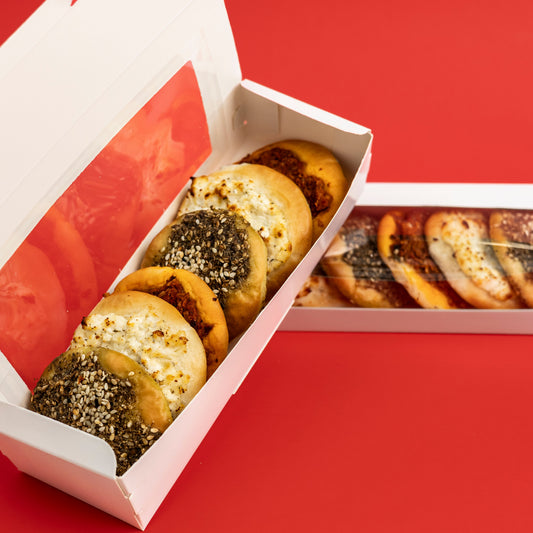 Two white cardboard bakery boxes containing assorted bagels and round baked goods, photographed on a solid red background. One box is open, showing the front bagels, and the other lies beside it showing the back side with a clear window.