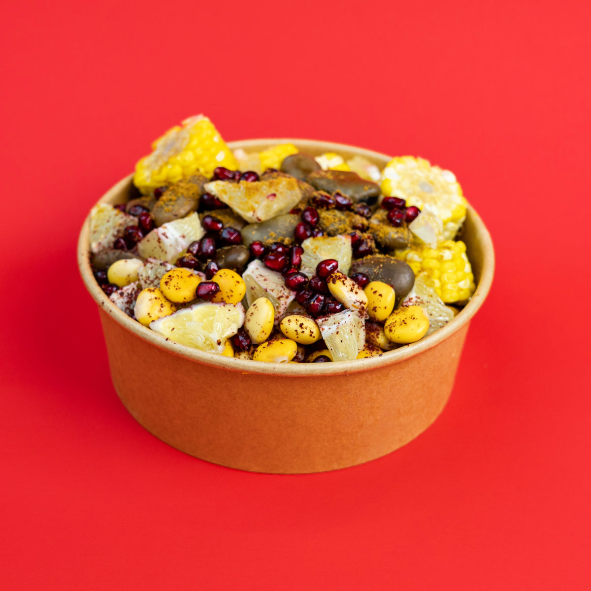 A paperboard bowl filled with a colorful snack mix of corn, beans, nuts, and dried fruit pieces, set against a solid bright red background.