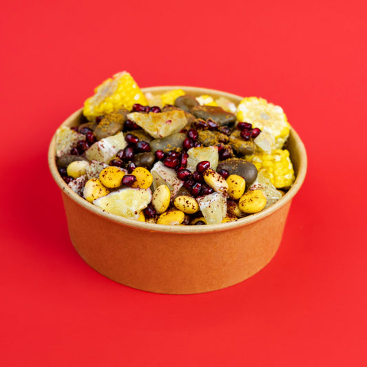 A paperboard bowl filled with a colorful snack mix of corn, beans, nuts, and dried fruit pieces, set against a solid bright red background.