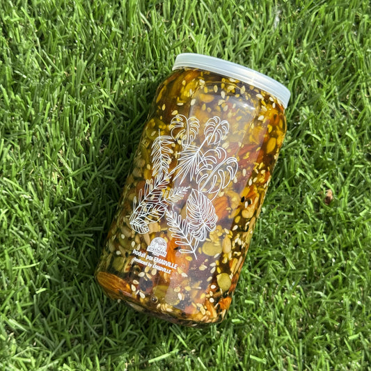 A clear glass jar filled with pickled vegetables and herbs, featuring a white lid and white printed illustrations of flowers and pine trees, lying on green grass.