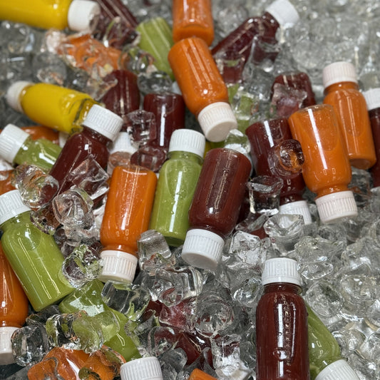 Small plastic bottles filled with orange, green, and dark red liquids, arranged on a bed of clear ice cubes.