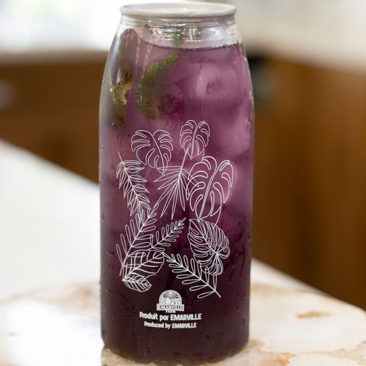 A clear glass can cooler filled with a dark purple iced drink, featuring white botanical line art and the text 'Peach & Wild Strawberry' on the front, resting on a light stone surface.