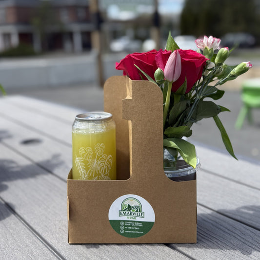 A brown cardboard carrier holding a glass can of yellow beverage and a small glass jar with pink and red flowers, placed on a gray picnic table outdoors with a blurred storefront background.