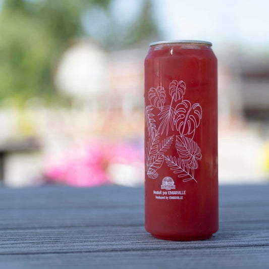 A red beverage can with white botanical line art and small text near the bottom, standing on a gray wooden surface with a blurred outdoor background.